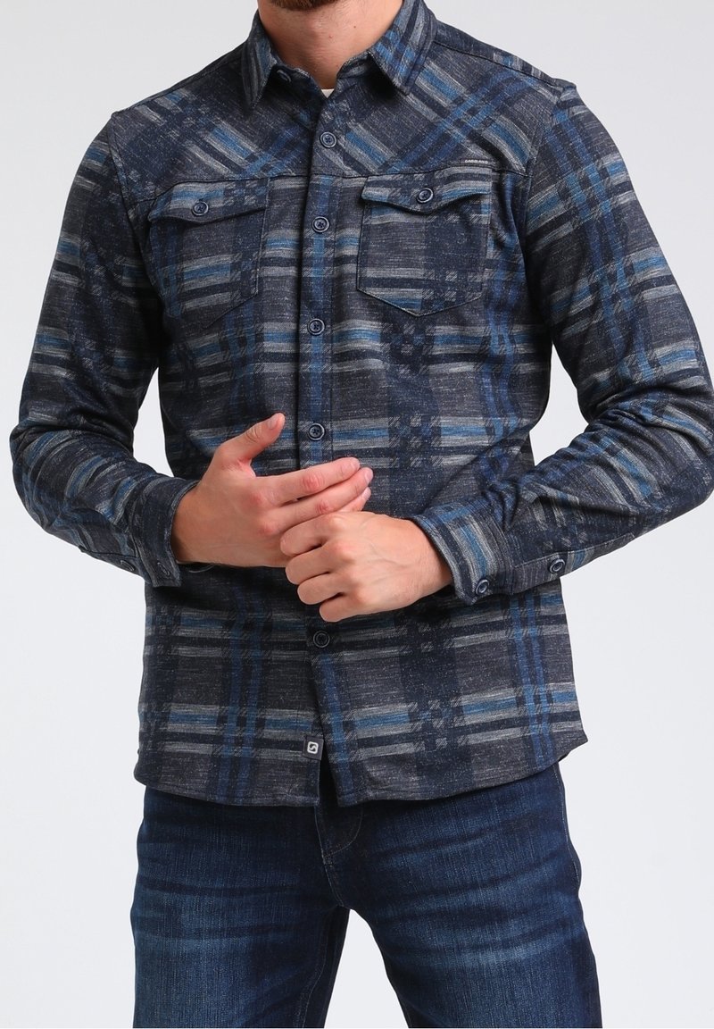 Homme portant une chemise à carreaux bleu foncé et grise à boutons avec deux poches poitrine et un jean bleu foncé, les mains jointes à la taille.
