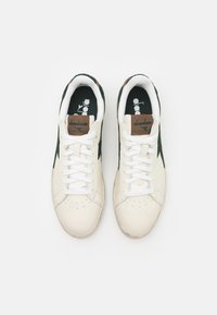 Hvite lær sneakers med grønne semskede detaljer, rund tå, hvite snørebånd og en brun logo-lapp på tungen. Strukturert gummisåle.