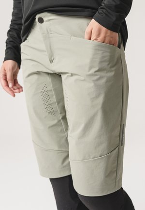 Person trägt hellgraue Shorts mit perforierter Belüftung an den Oberschenkeln und ein schwarzes langärmeliges Shirt, die Hand in der rechten Tasche.