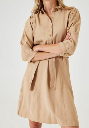 Femme portant une robe beige boutonnée avec des manches trois-quarts retroussées, croisant les bras devant un fond clair uni.