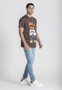 Bruine graphic t-shirt met een berenillustratie aan de voorkant, gecombineerd met lichtblauwe distressed jeans en witte sneakers.
