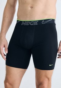 Boxers noirs Nike avec une ceinture logotée verte et noire. Tissu lisse, longueur mi-cuisse, design ajusté, sans motifs ni accents visibles.