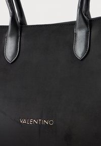 Crna kožna torba s glatkom teksturom, koja ima duple ručke i zlatni "VALENTINO" logo na donjem prednjem dijelu.