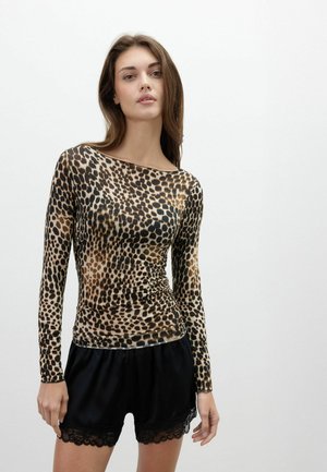 Ung kvinde iført en langærmet leopardprint top og sorte shorts med blondestrimler står foran en ensfarvet hvid baggrund.