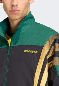Grön och svart träningsjacka med gula detaljer. Har en hög krage, dragkedja fram och Adidas-logotyp på bröstet. Texturerat tyg och ränder.