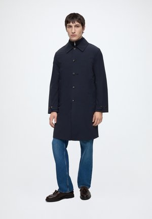 Mann steht und trägt einen langen dunkelblauen Mantel, blaue Jeans und dunkelbraune Lederschuhe vor einem schlichten hellen Hintergrund.