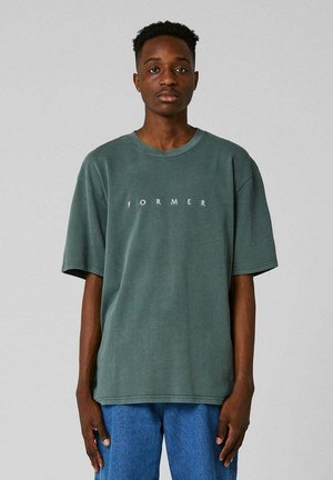 Jeune homme debout face à l’avant, portant un T-shirt vert avec le texte « FORMER » et un jean bleu sur un fond gris uni.