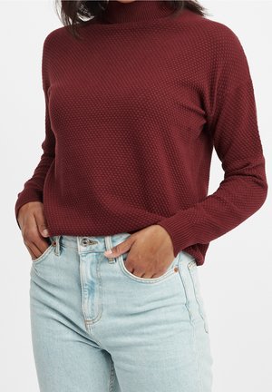 Frau trägt einen strukturierten weinroten Rollkragenpullover, der in hellblaue hochtaillierte Jeans gesteckt ist, mit Händen in den Taschen.