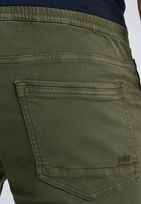 Olijfgroene broek met een gladde textuur, voorzien van achterzakken en contrasterende stiksels. Bevat een klein logodetail op de zak.