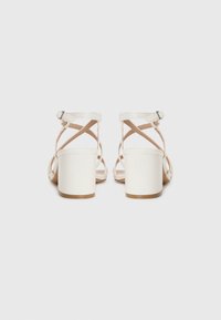 White heeled sandals with a block heel, featuring crisscross straps and a buckle closure at the ankle. Smooth texture and minimalist design.