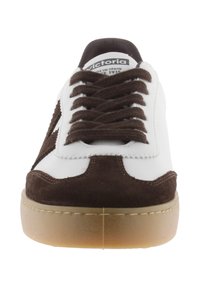 Deportiva blanca con acentos de ante marrón, con punta redonda, cordones planos y suela de goma marrón. Logo de la marca visible en la lengüeta.