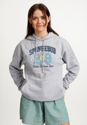 Ung kvinde iført grå SpongeBob-hoodie med nummer 99 og grøn-hvide stribede shorts, står med hænderne i lommerne på hvid baggrund.