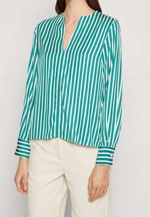 Kvinde iført en langærmet bluse med lodrette striber i grønblå og hvid med manchetknapper og cremefarvede bukser.