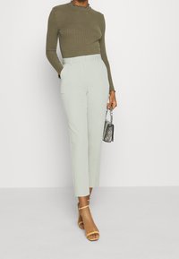 Topp med rysjer i olivengrønn, langermet, kombinert med høytlivte lysegrønne bukser og beige sandal med stropper.