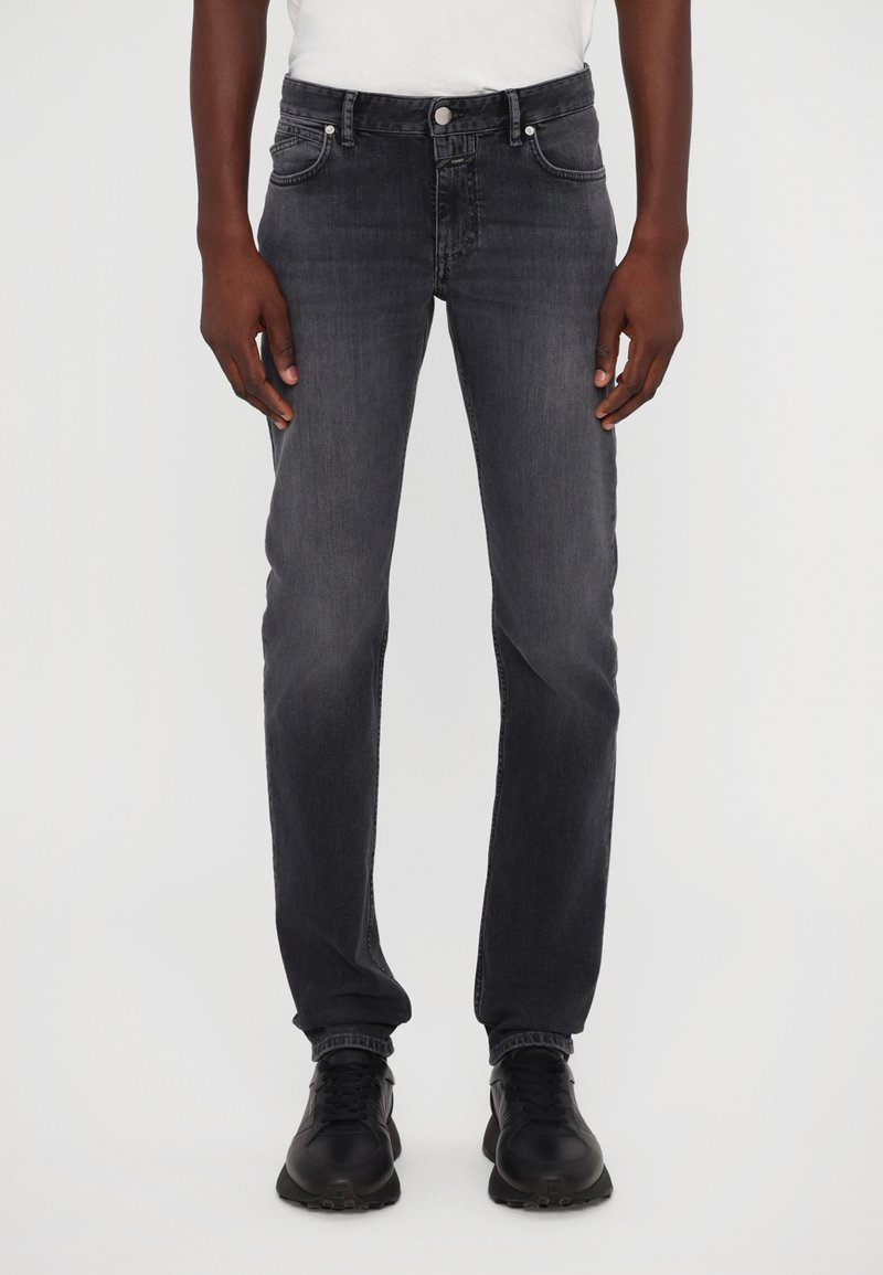 Jeans de mezclilla gris oscuro con corte recto, bolsillos frontales y cierre de botón. Llevados con zapatillas negras para crear contraste.