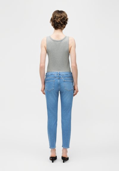 Personne aux cheveux courts et bouclés portant un haut gris sans manches, un jean bleu clair et des talons ouverts noirs, debout de dos.