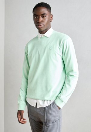 Mint green sweatshirt with crew neckline, ribbed cuffs, and embroidered logo. Paired with a white collared shirt and striped pants.