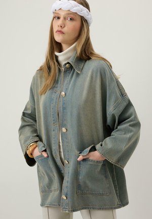 Młoda kobieta w oversize'owej wypłowiałej dżinsowej kurtce, białym golfie, białej plecionej opasce na głowę i złotych bransoletkach, z rękami w kieszeniach kurtki.