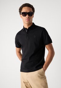 Črna polo majica s ovratnikom in kratkimi rokavi, ki ima diskreten logotip na prsih. Oblečena je s svetlim bež hlačami in temnimi sončnimi očali.