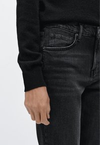 Černý pletený svetr s žebrovaným lemem, nošený přes černé džíny s rovnými nohavicemi. Džíny mají mírný lesk a klasický pětikapsový střih.