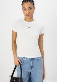Camiseta de algodón blanca con mangas cortas, presentando un diseño de logo circular en rojo y azul en el pecho. Combinada con unos jeans azules y un bolso negro.
