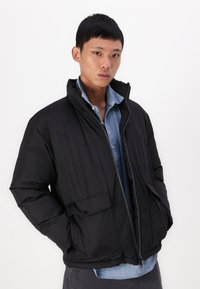 Jachetă puffer neagră cu guler înalt, fermoar frontal și două buzunare laterale. Purtată peste o cămașă din denim albastru deschis, cu o textură netedă.