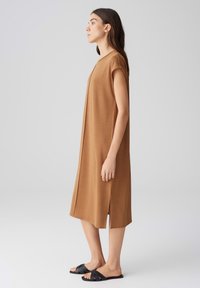 Braunes knielanges Kleid mit kurzen Ärmeln, seitlichen Schlitzen und glatter Textur, kombiniert mit schwarzen Riemchensandalen. Minimalistisches Design.