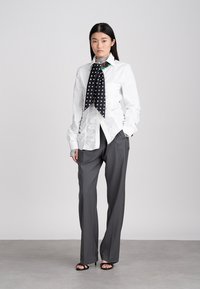 Γυναίκα με λευκό μπλουζάκι με ρυτίδες, μαύρο φουλάρι με πουά σχέδιο, γκρι παντελόνι φαρδύ και μαύρες γόβες ανοιχτού τύπου να κάθεται σε γκρι δάπεδο.