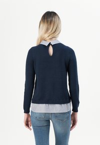 Pull en maille bleu marine avec une ouverture en forme de goutte au dos, accompagné d'une chemise à col rayé en dessous. Associé à un jean bleu pour un look superposé.