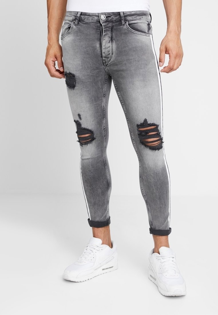 Jeans de mezclilla gris entallados con detalles desgastados y franjas blancas a los lados, que incluyen una cremallera y puños enrollados.