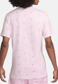 T-shirt Nike rose clair avec un motif de logo répétitif et du texte. Manches courtes et coupe décontractée. Design simple à col rond.