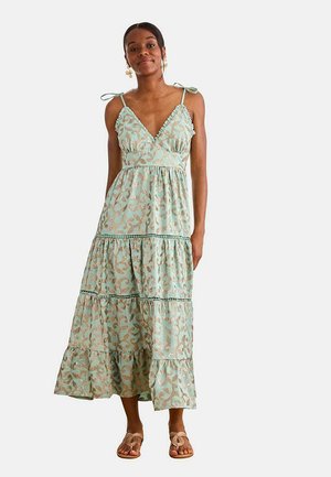 Mujer con un vestido maxi sin mangas, estampado, verde menta, con tirantes de lazo y falda de volantes, de pie con postura relajada y sandalias.