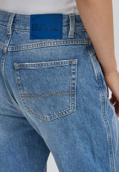 Ljust blå denimjeans med en bakficka som har kontrasterande bruna sömmar. En blå läderlapp med texten "BALL" är fäst vid midjan.