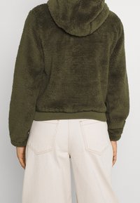 Oliwkowa polarowa kurtka z kapturem, ribbowanym dołem i mankietami. Materiał ma miękką, teksturowaną powierzchnię. Noszona na jasnych spodniach.