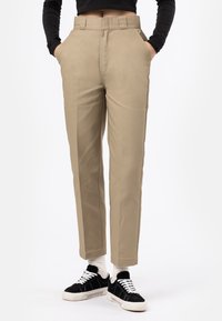Pantalon beige sur mesure à coupe droite, équipé de poches latérales et d'une finition lisse, associé à des baskets noires et des chaussettes blanches.