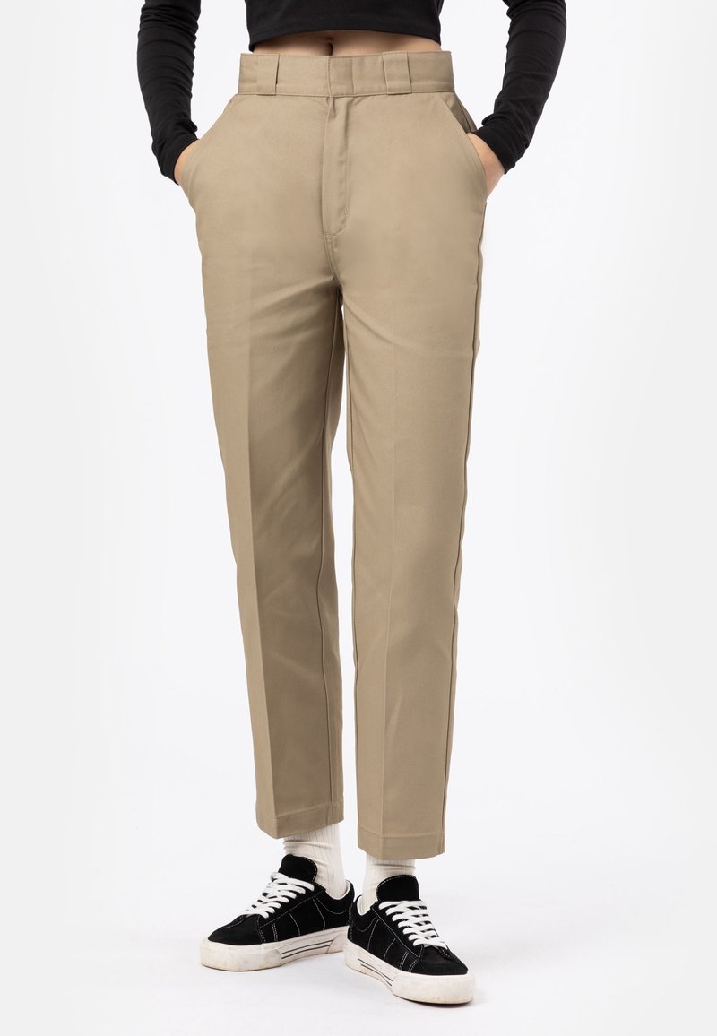 Pantalon beige sur mesure à coupe droite, équipé de poches latérales et d'une finition lisse, associé à des baskets noires et des chaussettes blanches.