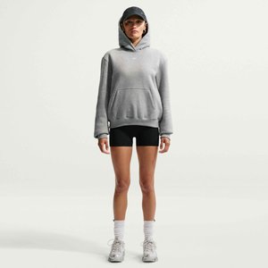 Donna con felpa grigia con cappuccio, berretto nero, pantaloncini da ciclismo neri, calzini bianchi e sneakers grigie, in piedi davanti a uno sfondo chiaro e semplice.