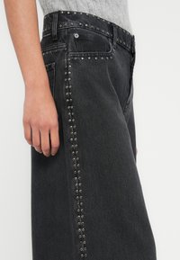 Μαύρα τζιν denim με ασημένια διακοσμητικά καρφάκια κατά μήκος της πλαϊνής ραφής, με κλασική μέση και χαλαρή εφαρμογή.