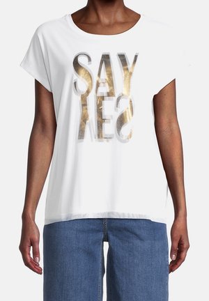 Persoon draagt een wit shirt met korte mouwen met reflecterende gouden en grijze tekst "SAY YES" en een blauwe spijkerbroek.