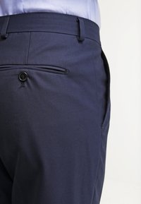 Navy blue trousers made of textured fabric, featuring a rear pocket with a black button and single belt loop. Clean, tailored design.