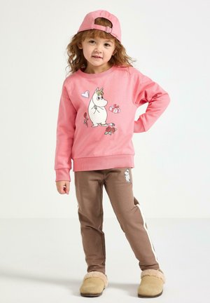 Rosa sweatshirt med karaktärstryck, kombinerad med bruna byxor med accent och ljusa sidostripes. Beige fluffiga tofflor kompletterar outfiten.