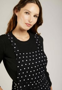 Robe noire à pois blancs, avec des bretelles réglables, portée par-dessus un pull côtelé noir. Texture lisse, design ajusté.