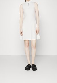 Weißes, ärmelloses Kleid mit hohem Kragen und einem Reißverschluss vorne. Ausgestelltes Design, kombiniert mit schwarzen Plateauschuhen mit einer Schnallenverzierung.
