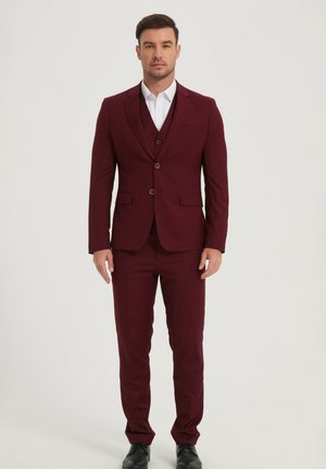 Homme debout portant un costume bordeaux ajusté avec une chemise blanche et des chaussures noires, sur un fond clair uni.