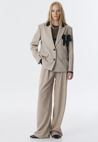 Beige op maat gemaakte blazer met zwarte revers, gecombineerd met een broek met brede pijpen. Heeft een textuurpatroon en een decoratieve zwarte strik op de zak.