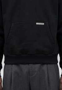 Sweat-shirt noir avec poche kangourou devant et petite étiquette rectangulaire, associé à un pantalon plissé gris.