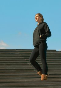 Schwarzer Hoodie mit einer Vordertasche und Logo, kombiniert mit schwarzen Leggings und braunen Knöchelstiefeln, steht auf Holzstufen vor einem blauen Himmel.