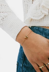 Pulsera de mariposa dorada en la muñeca; cuenta con una cadena delicada y un diseño de mariposa brillante y simétrico. Se lleva con un suéter blanco texturizado.