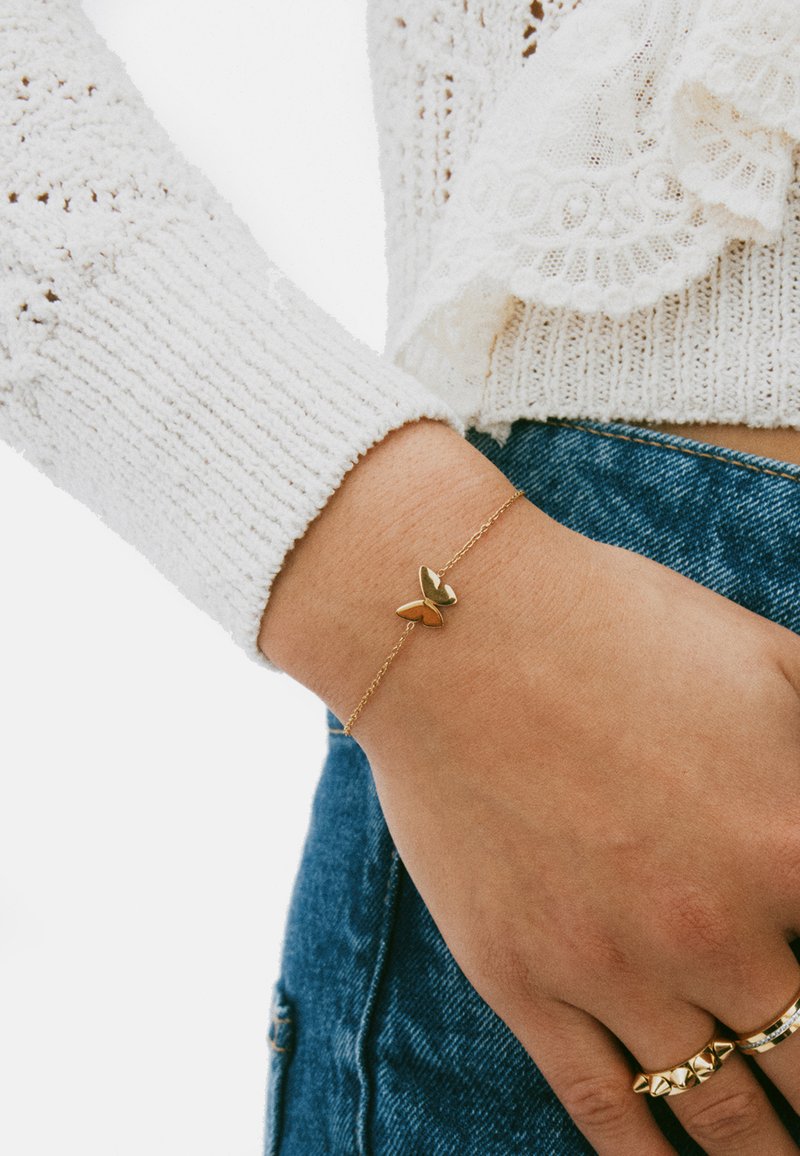 Pulsera de mariposa dorada en la muñeca; cuenta con una cadena delicada y un diseño de mariposa brillante y simétrico. Se lleva con un suéter blanco texturizado.