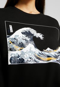Sweatshirt noir avec un motif imprimé de grandes vagues aux accents bleus et blancs, un petit bateau et des silhouettes de montagnes en arrière-plan.